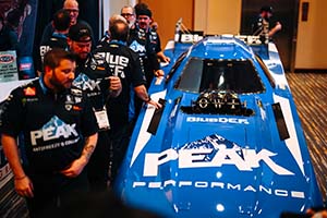 Blue and white Funny Car with Peak Performance logo and men wearing black shirts and pants on left