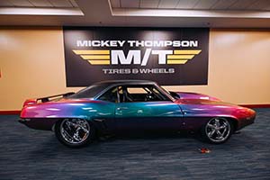 Camaro with paint that transitions from red to purple to blue to green under black Mickey Thompson M/T Tires & Wheels sign